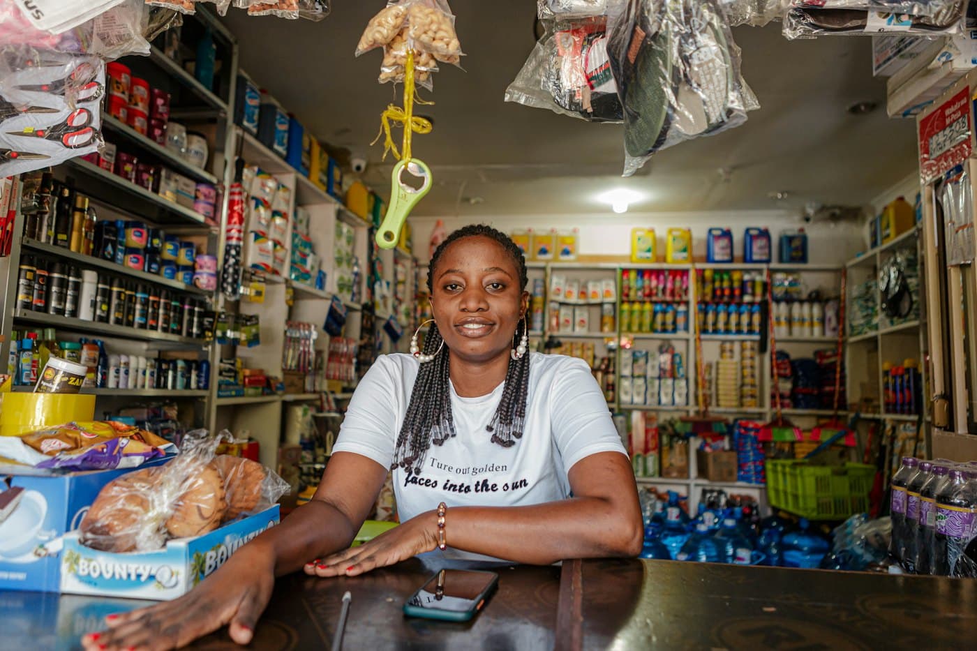 A shopkeeper smiling behind the counter of her provisions shop, shelves stocked behind her and her phone on the counter.